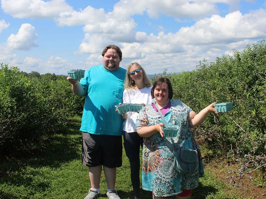 Three people in a blueberry field holding up boxes of freshly picked berries