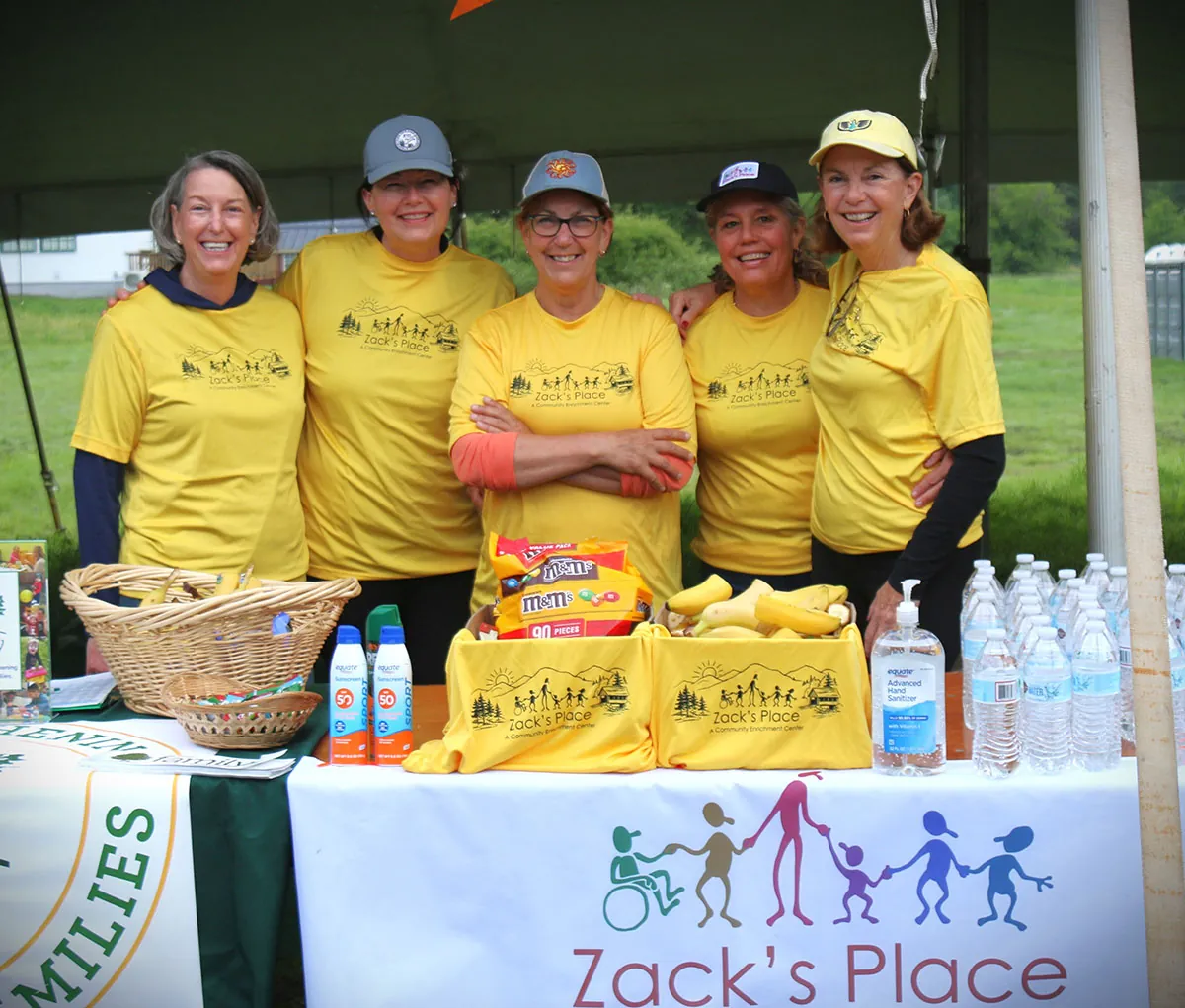 Five smiling women in yellow Zack’s Place T-shirts at the Zack’s Place table at an outdoor event, with snacks, sunscreen and bottled water